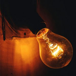 A close-up view of an illuminated incandescent light bulb glowing warmly against a dark background.